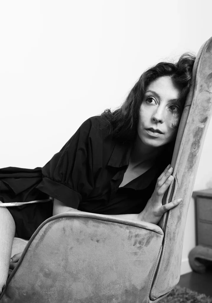 Une femme brune vêtue de noir s’appuie au dossier d’un fauteuil en velours, en noir et blanc.
