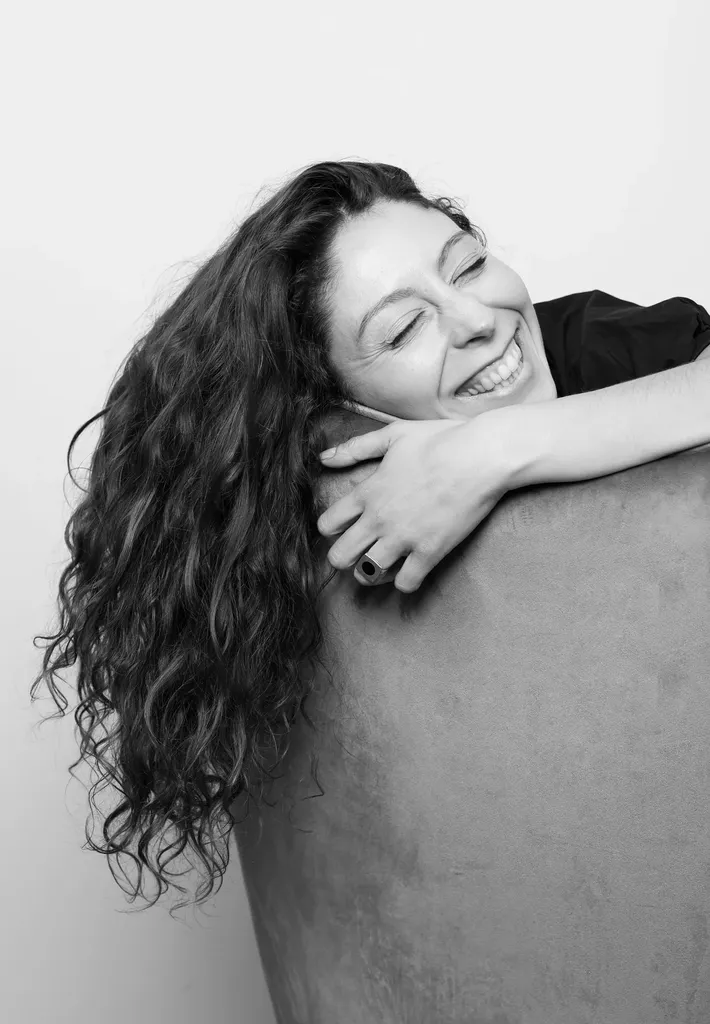 Une femme aux cheveux bouclés rit, les bras posés sur le dossier d’un fauteuil, en noir et blanc.