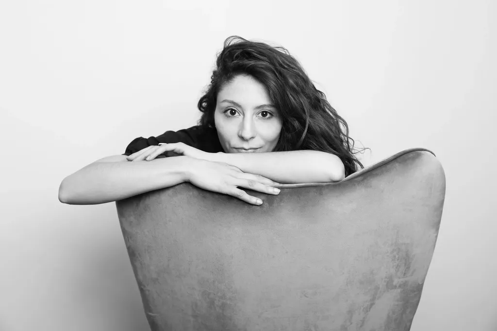 Une femme aux cheveux bouclés pose le menton sur ses bras croisés au dossier d’un fauteuil, cadrage horizontal en noir et blanc.