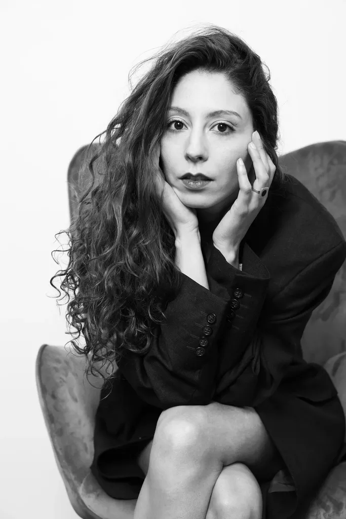 Une femme aux cheveux bouclés assise dans un fauteuil, les mains près du visage, en noir et blanc.