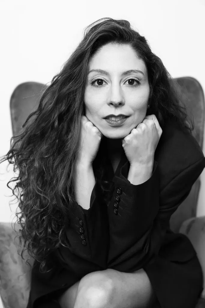 Une femme aux cheveux bouclés assise dans un fauteuil, regard direct, poings sous le menton, en noir et blanc.