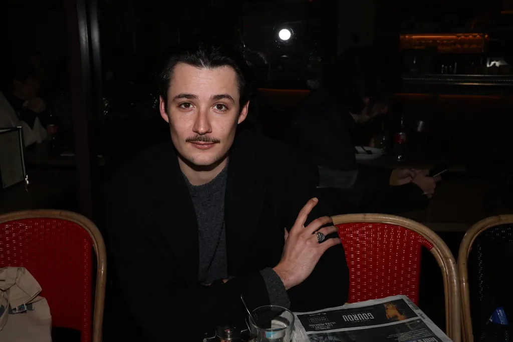Un jeune homme moustachu assis à une terrasse de café le soir, entre des chaises rouges, cadrage horizontal.