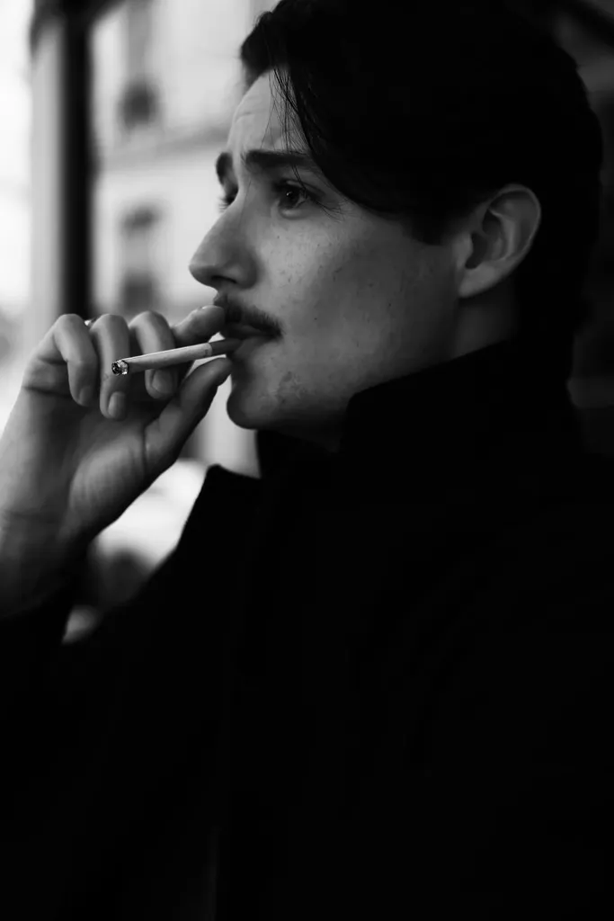 Profil d’un jeune homme moustachu, une cigarette aux lèvres, fort contraste en noir et blanc.
