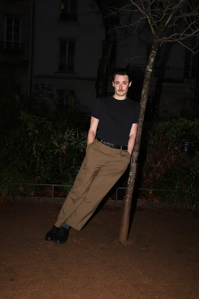 Un jeune homme en t-shirt noir adossé à un arbre, de nuit, devant des immeubles.