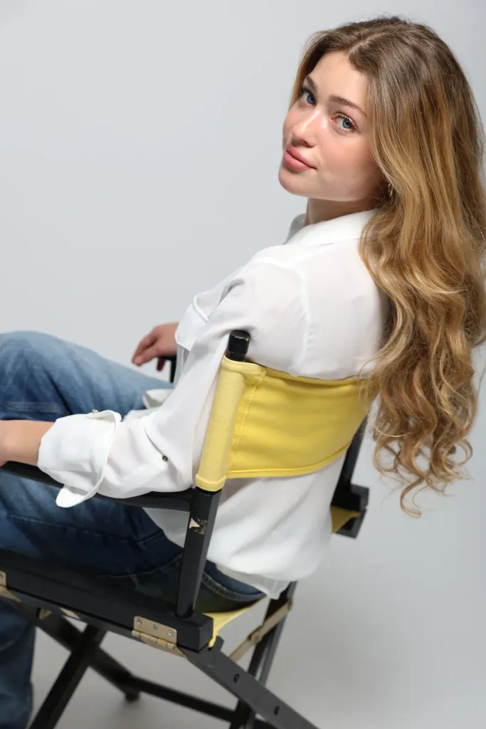 Une jeune femme en chemise blanche assise de profil sur une chaise de réalisateur à toile jaune, regard vers l’objectif.