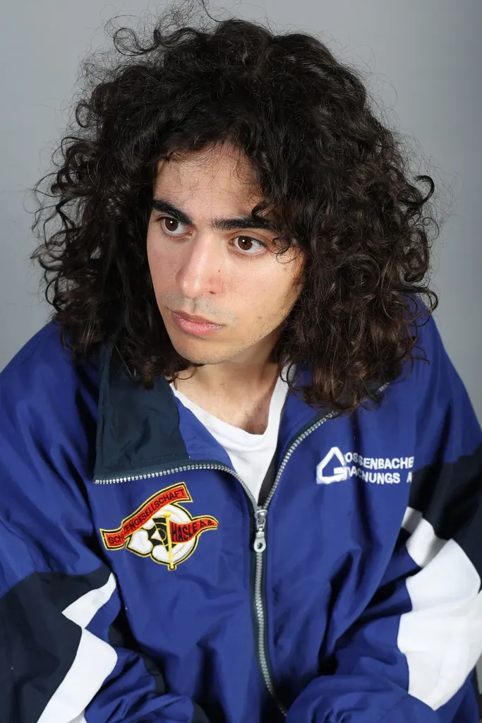 Portrait d’un jeune homme aux cheveux bouclés en veste de survêtement bleue, fond gris.
