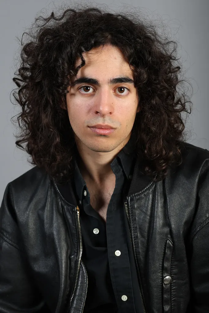 Portrait de face d’un jeune homme aux cheveux bouclés en veste de cuir noire, fond gris.