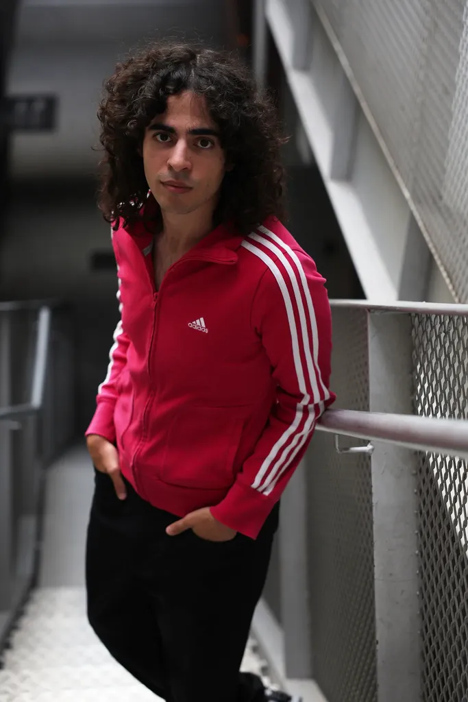 Un jeune homme aux cheveux bouclés en veste rose, les mains dans les poches, dans un escalier métallique.