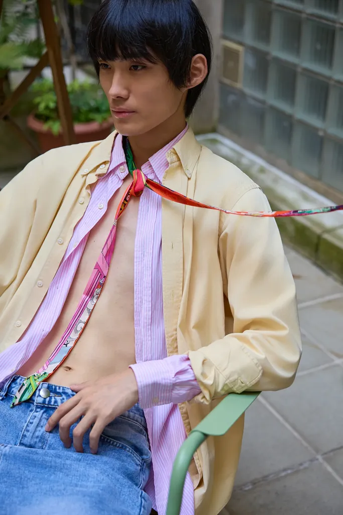 Le même jeune homme assis sur une chaise verte, chemise jaune sur chemise rayée rose et foulard coloré, cadré de près devant des briques de verre, en couleur.