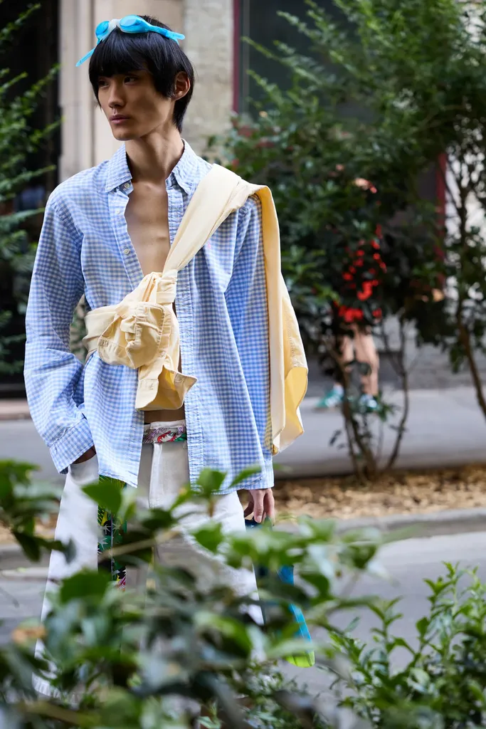 Le même jeune homme en chemise vichy bleue, un pull jaune noué à la taille, entouré de feuillage dans une rue, en couleur.