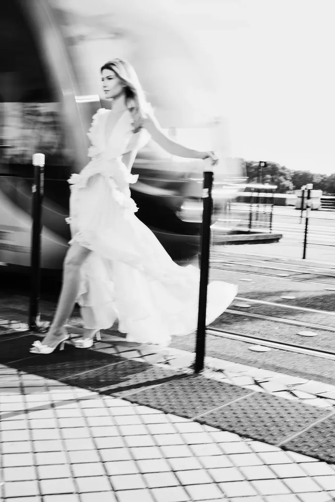 Femme en longue robe blanche à volants marchant sur des pavés, l’arrière-plan filé par le mouvement, en noir et blanc.
