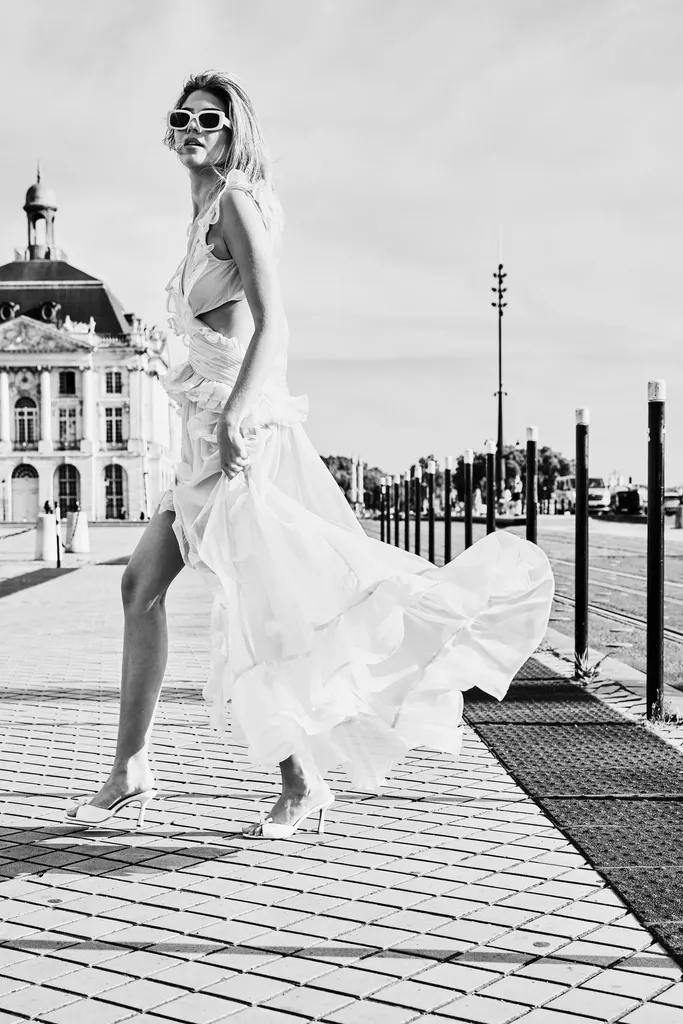 La même femme en robe blanche à volants et lunettes blanches, la robe soulevée par le pas, sur un quai pavé bordé de bornes, en noir et blanc.