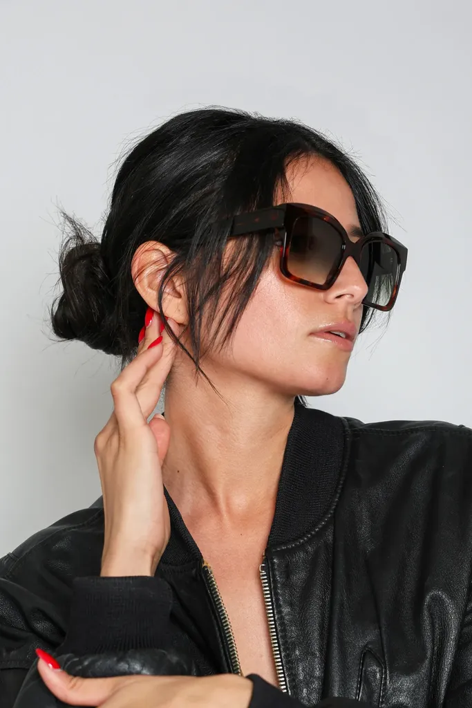 Portrait serré d’une femme brune en grandes lunettes de soleil et blouson de cuir noir, une main à l’oreille, sur fond gris de studio, en couleur.