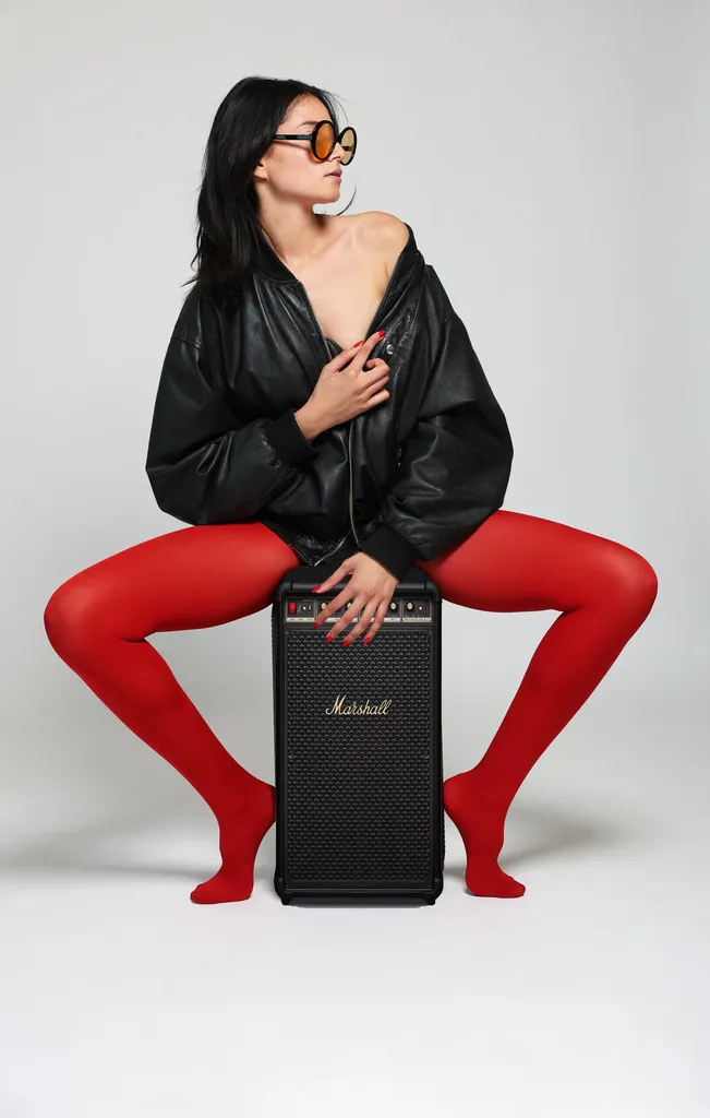 La même femme en blouson de cuir noir et collants rouges, assise sur un ampli noir, lunettes rondes orangées, sur fond clair de studio, en couleur.