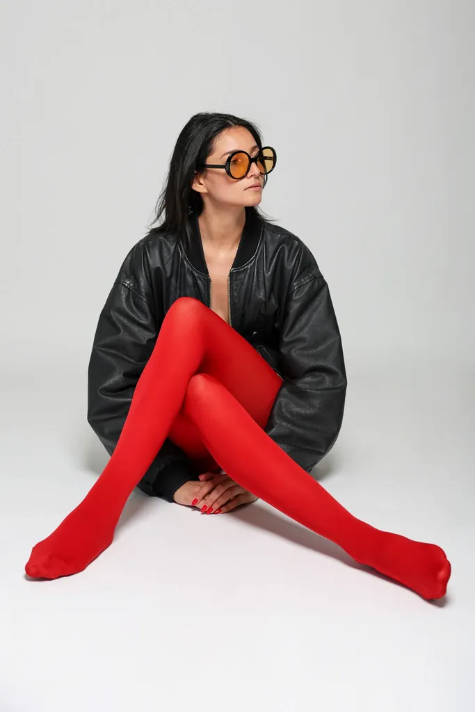 La même femme assise au sol, jambes en collants rouges allongées, blouson de cuir noir, sur fond clair de studio, en couleur.