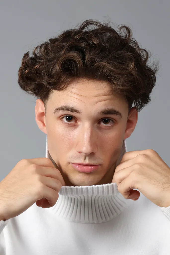 Gros plan d’un jeune homme aux cheveux bouclés qui remonte le col d’un pull blanc à col roulé, sur fond gris, en couleur.