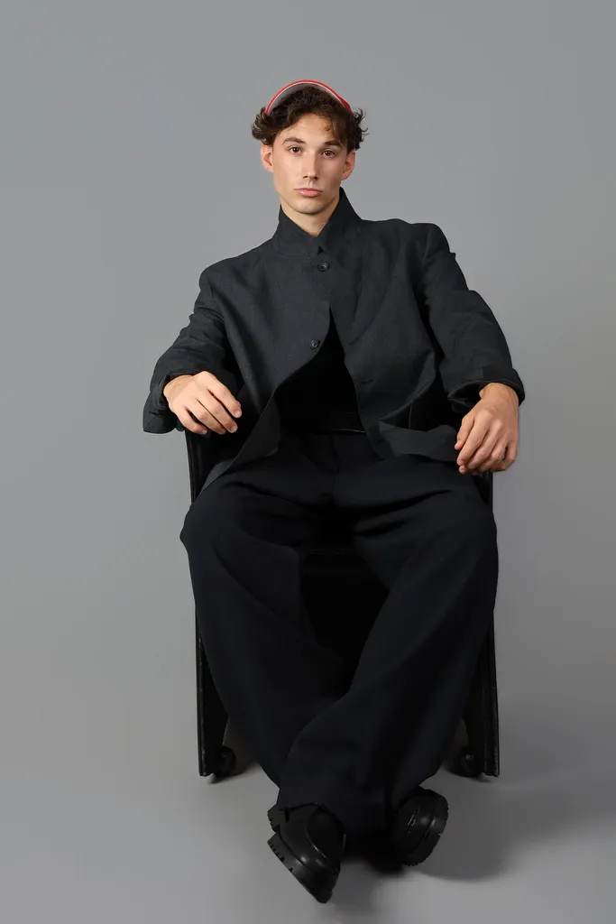 Le même jeune homme assis en costume noir et casquette rouge, en pied sur fond gris de studio, en couleur.