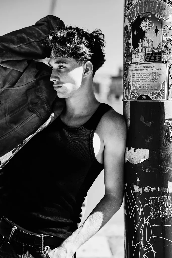 Jeune homme en débardeur noir et blouson de cuir adossé à un poteau couvert d’affiches, en plein soleil, en noir et blanc contrasté.
