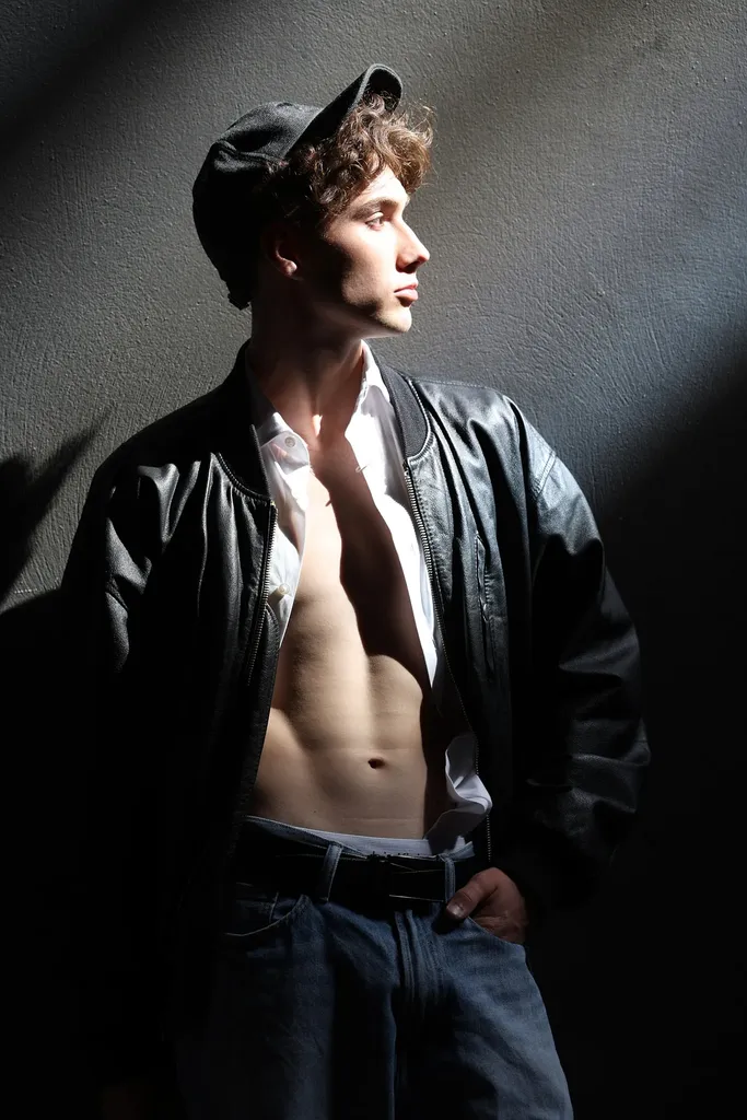 Jeune homme en casquette et blouson de cuir ouvert, torse nu, dans une lumière latérale dure contre un mur sombre, en couleur.