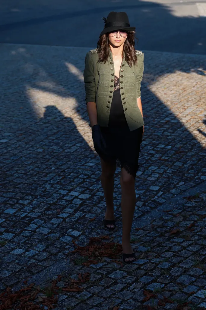 Femme en chapeau noir, veste kaki à boutons dorés et robe noire marchant sur des pavés dans une lumière rasante aux longues ombres, en couleur.