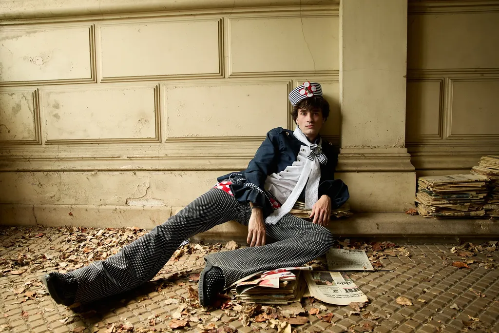 Jeune homme en veste bleu marine et pantalon rayé assis par terre contre un mur de pierre clair, parmi des feuilles mortes et des journaux, en couleur.