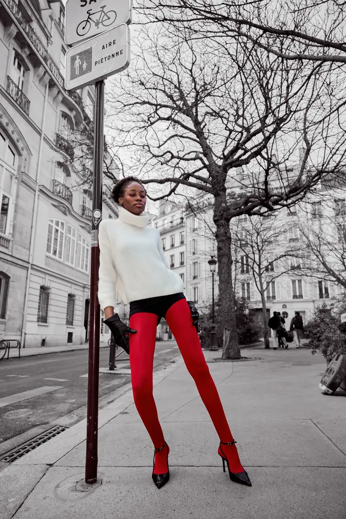Femme en pull blanc à col roulé et collants rouges appuyée à un poteau sur un trottoir, la rue en noir et blanc et les collants en couleur.
