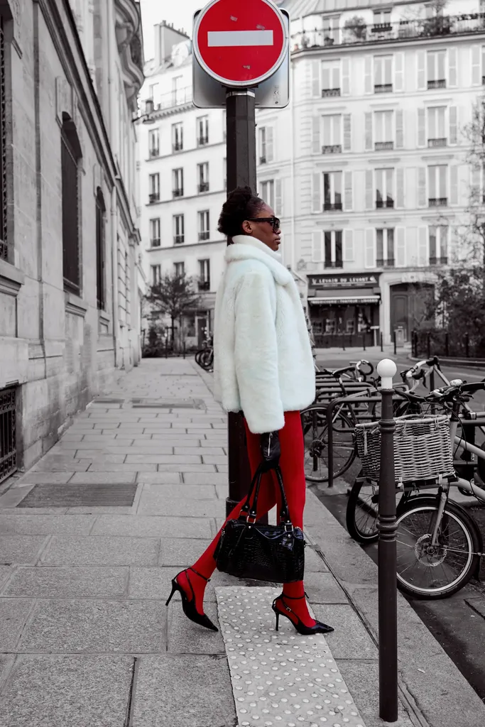 Femme en manteau de fourrure blanche, lunettes noires et collants rouges sur un trottoir près de vélos, la rue en noir et blanc et les collants en couleur.