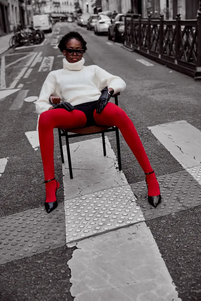 Femme assise sur une chaise au milieu d’un passage piéton, pull blanc, gants noirs et collants rouges, la rue en noir et blanc et les collants en couleur.