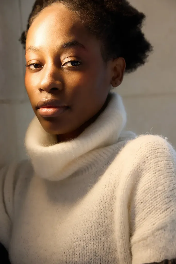 Portrait serré d’une jeune femme en pull crème à col roulé, éclairée par une lumière chaude de côté, en couleur.