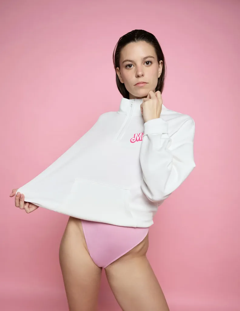 Femme aux cheveux courts en sweat blanc et body rose, tenant le bas de son sweat, sur fond papier rose, en couleur.