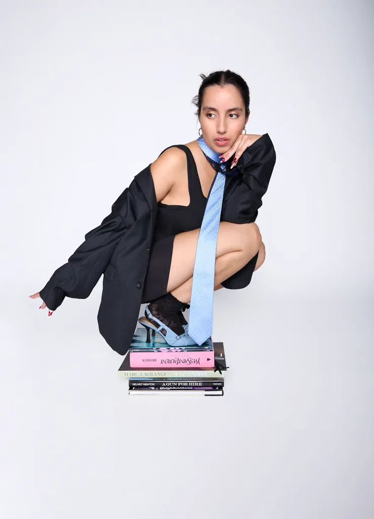 Femme accroupie sur une pile de livres, blazer noir trop grand et cravate bleu ciel, sur fond gris clair, en couleur.
