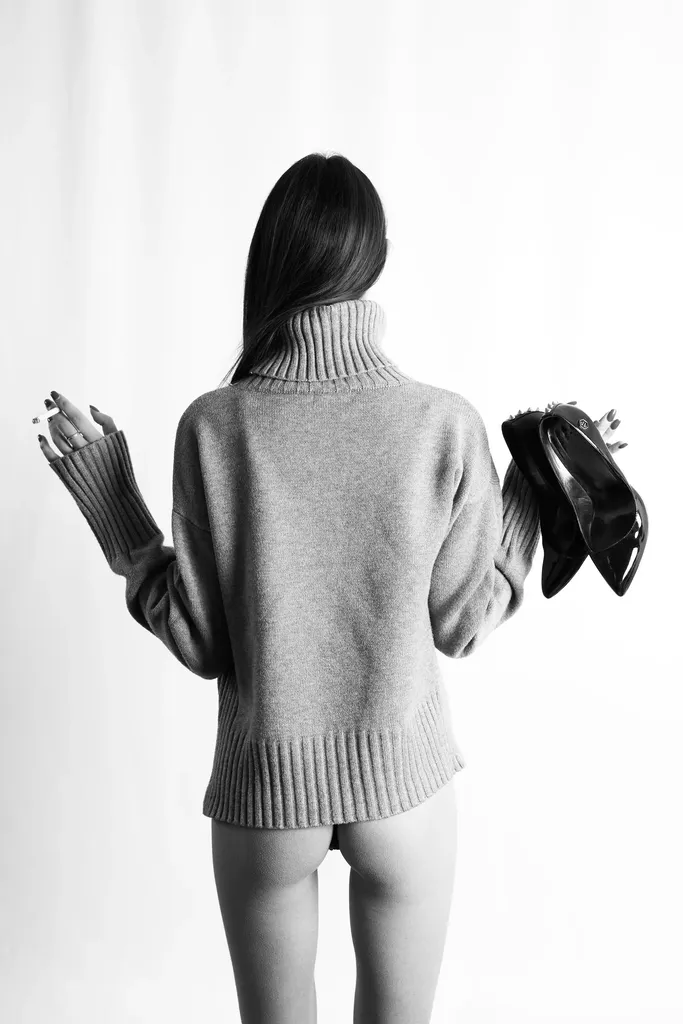 Femme de dos en pull à col roulé, une paire d’escarpins noirs à la main, sur fond blanc, en noir et blanc.