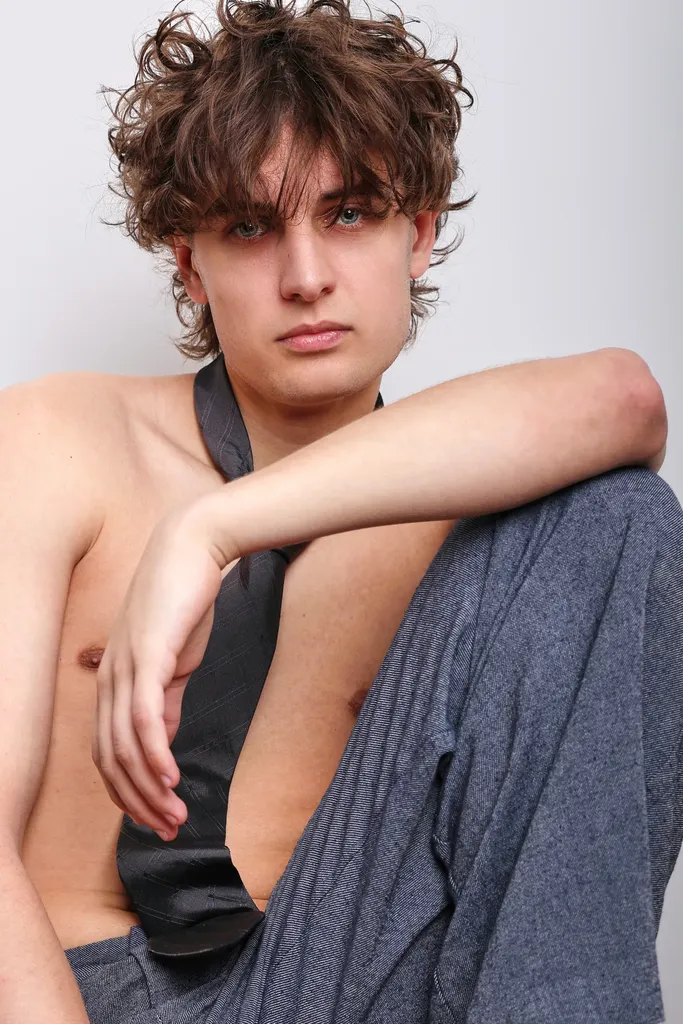 Jeune homme aux cheveux bruns ébouriffés, torse nu avec une cravate sombre, le bras posé sur un genou relevé, sur fond clair, en couleur.