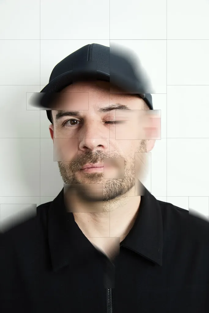 Portrait en couleur d’un homme en casquette et chemise noires face à l’objectif, le visage recomposé en carreaux décalés et flous sur un fond blanc quadrillé.