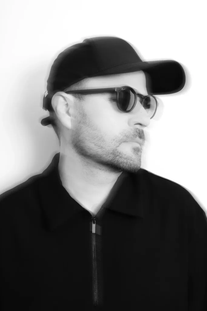 Portrait en noir et blanc d’un homme en casquette et lunettes de soleil tourné de profil vers la droite, les contours doublés par un flou de bougé, sur fond blanc.