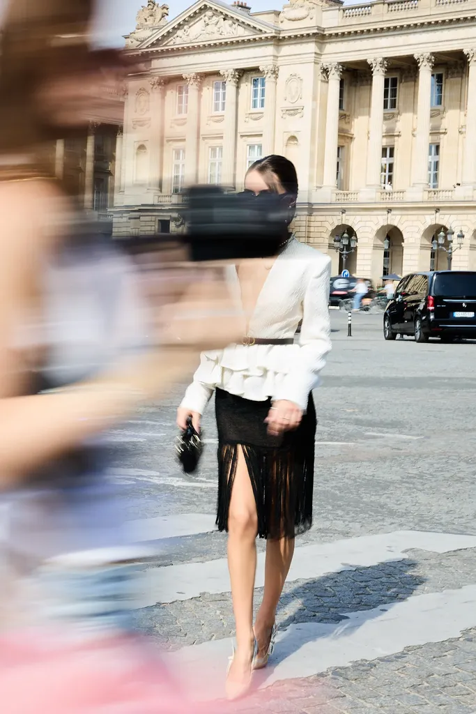 Couleur, dans la rue : une femme en veste blanche à volants et jupe noire à franges traverse un passage piéton, entre des silhouettes floues au premier plan.