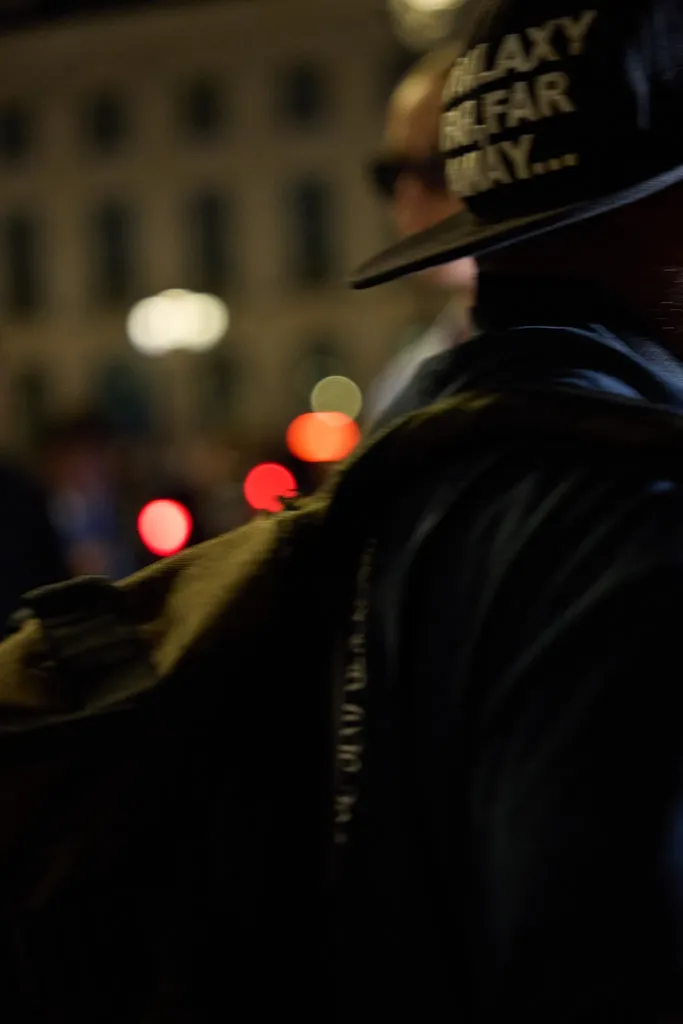 Couleur, de nuit : de dos, une personne en veste sombre et casquette brodée, devant les lumières floues d’une rue.