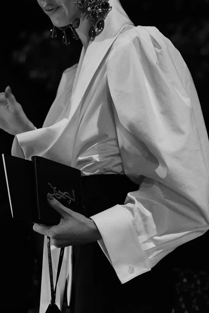 Noir et blanc : le buste d’une personne en chemise blanche ample à grandes manches, tenant une petite pochette noire, boucles d’oreilles pendantes.
