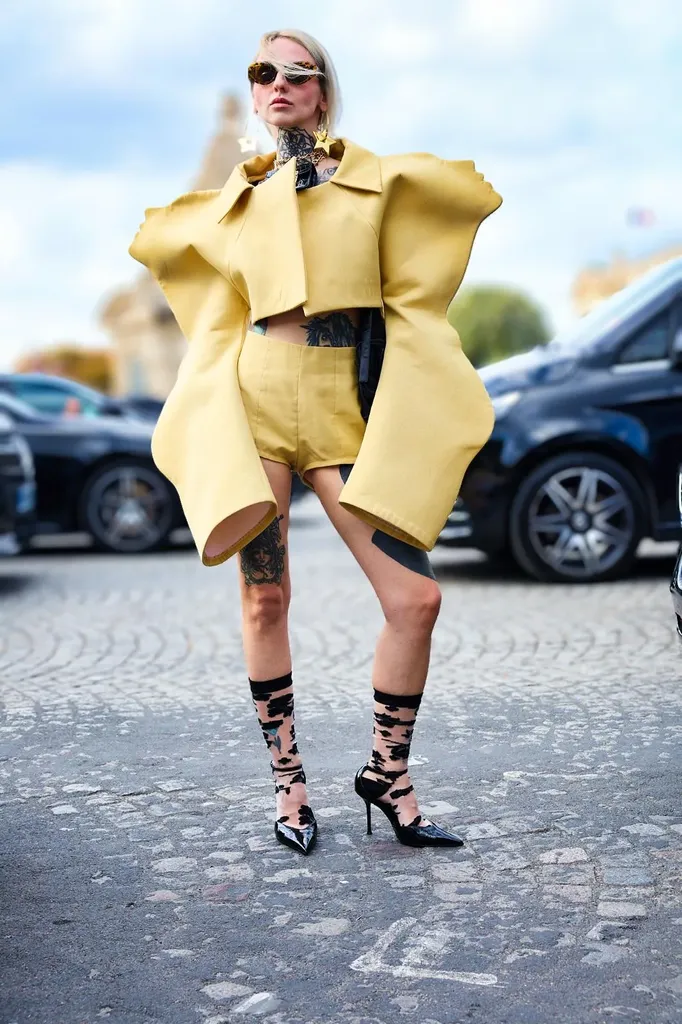 Couleur, en pied dans la rue : une femme en ensemble jaune aux épaules sculptées, chaussettes noires et escarpins, devant des voitures.