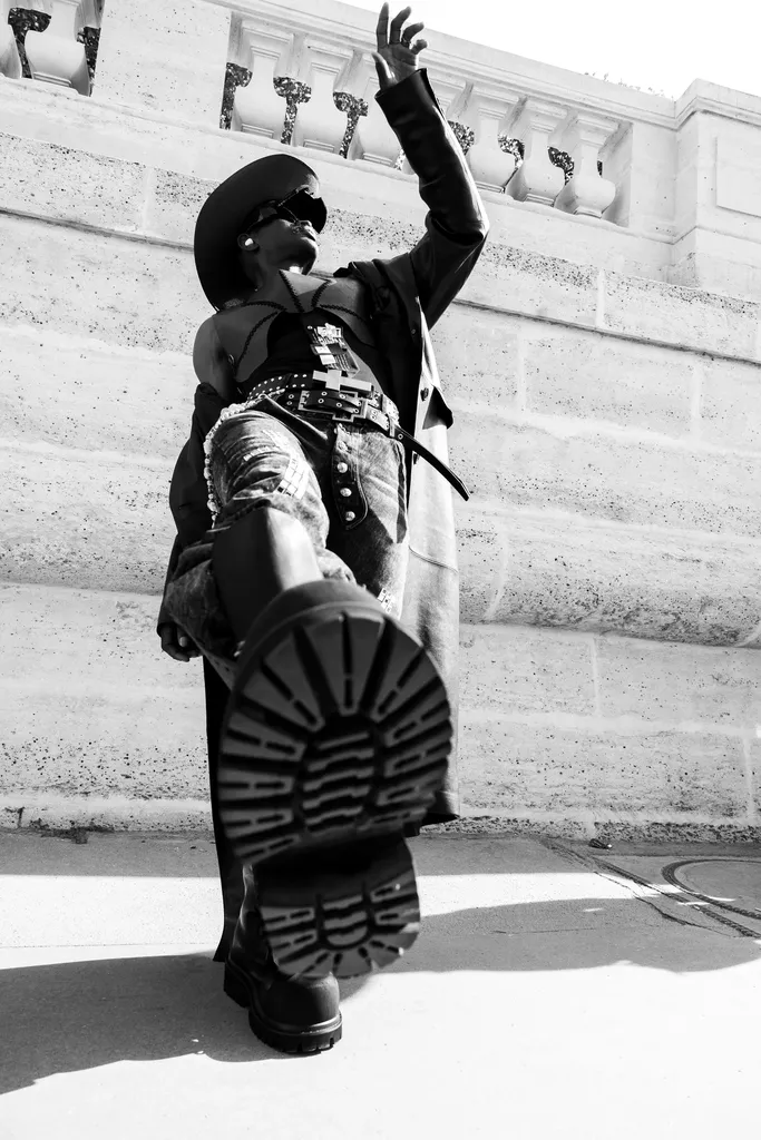 Noir et blanc, contre-plongée : une personne en chapeau et grosses bottes lève une semelle crantée vers l’objectif, devant un mur de pierre.