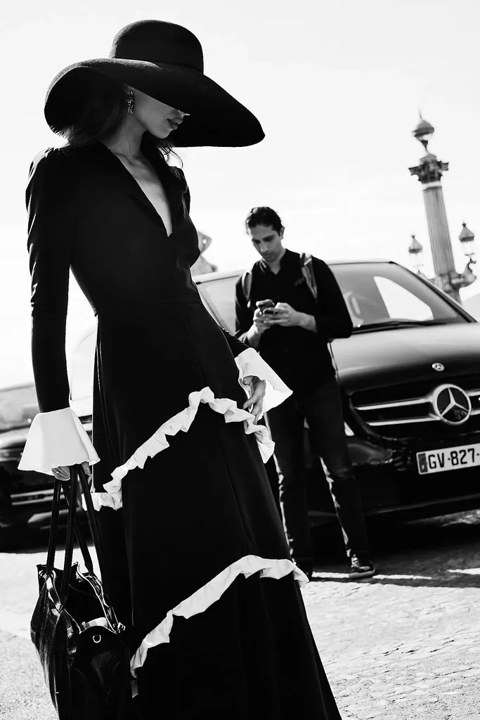 Noir et blanc, plein soleil : une femme en longue robe noire à volants blancs et chapeau à larges bords, devant une voiture noire.