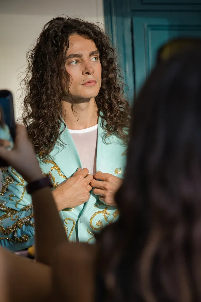 Couleur : un jeune homme aux longs cheveux bouclés, en veste turquoise brodée d’or, entouré de téléphones qui le photographient.