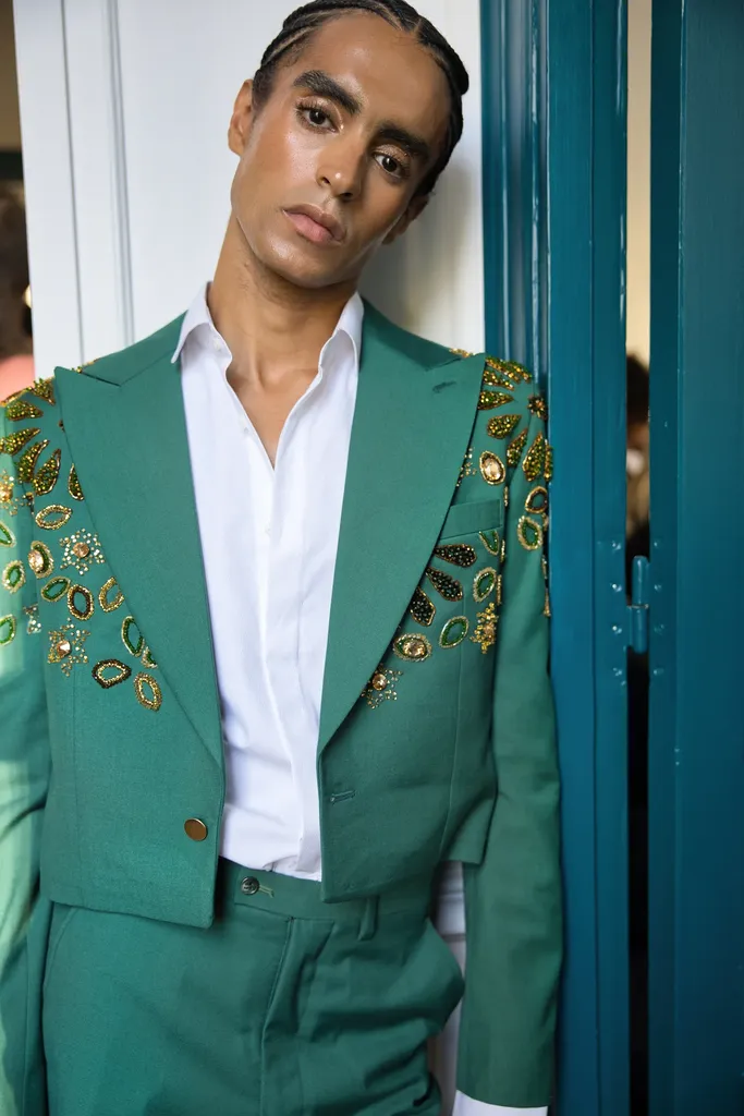 Couleur, portrait à mi-corps : un homme en costume vert brodé de sequins, chemise blanche ouverte, appuyé contre une porte bleu canard.