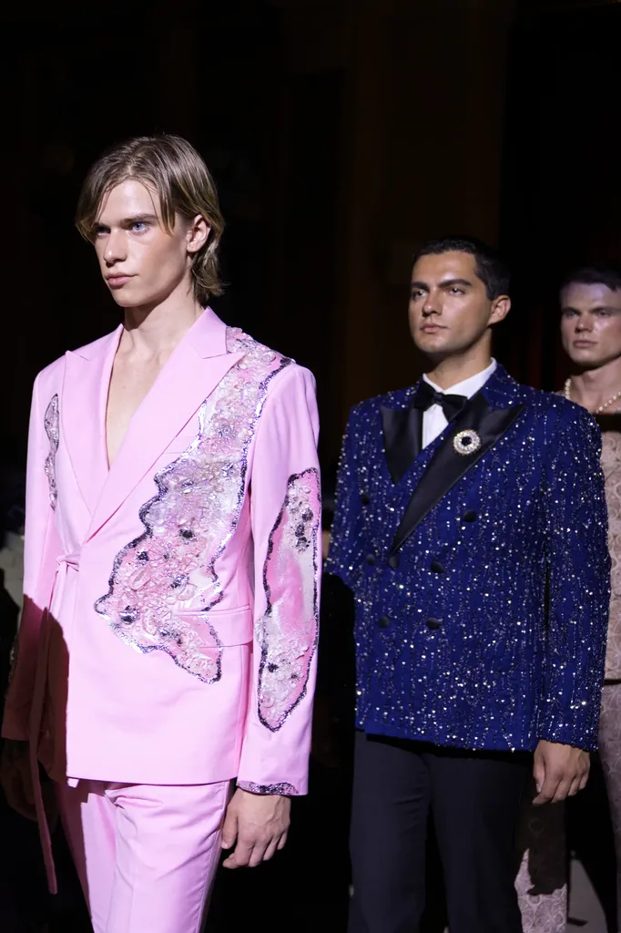 Couleur, sur un podium sombre : deux hommes en costume, l’un rose brodé, l’autre bleu pailleté avec nœud papillon.