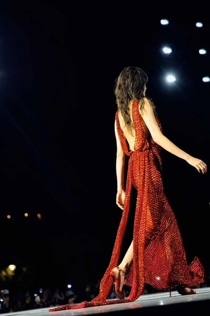 Couleur, défilé de nuit : de dos, une mannequin en longue robe rouge scintillante, dos nu, marche vers les projecteurs.