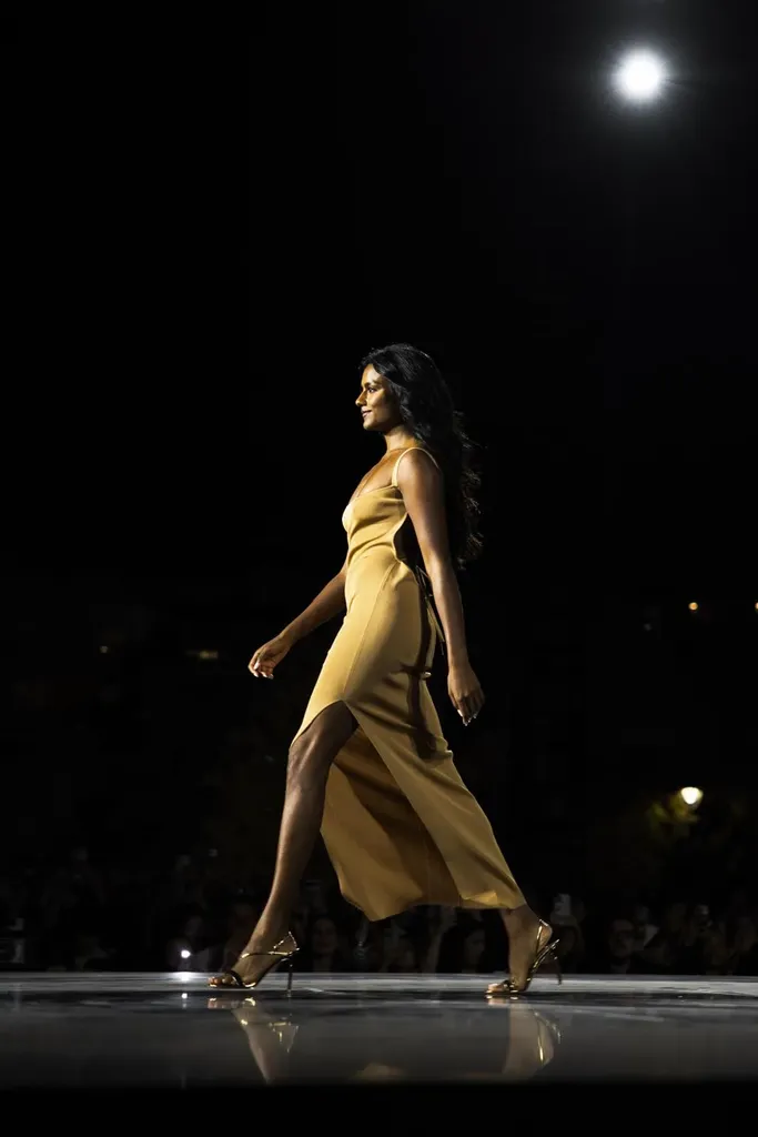 Couleur, défilé de nuit : une mannequin en robe de satin dorée marche sur un podium brillant, un projecteur au-dessus d’elle.