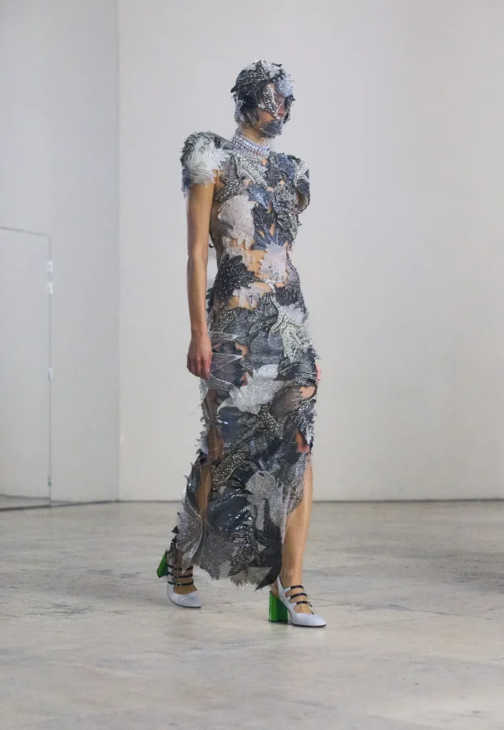 Couleur, présentation en salle claire : une mannequin en robe longue couverte de pièces argentées et grises, visage masqué.