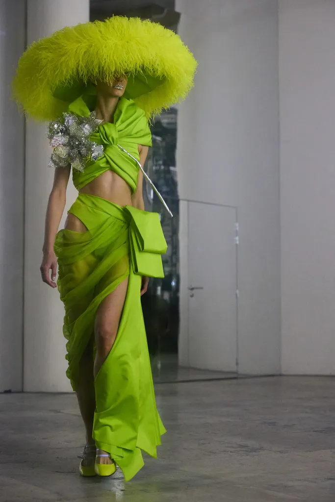 Couleur, présentation en salle claire : une mannequin en ensemble vert anis drapé, coiffée d’un immense chapeau en plumes.