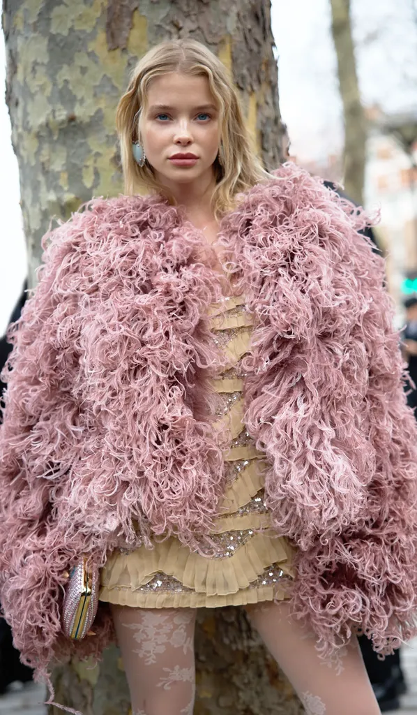 Couleur, dans la rue : une jeune femme blonde en manteau de plumes rose poudré et robe dorée, devant un tronc d’arbre.
