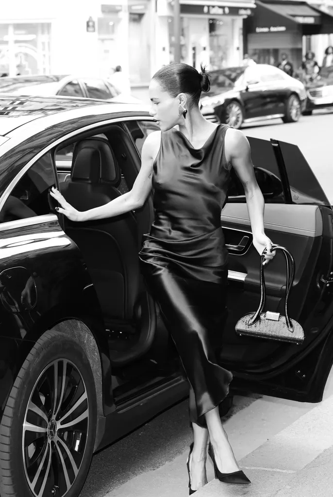 Noir et blanc : une femme en robe de satin sombre sort d’une voiture, la portière ouverte, sac à la main.
